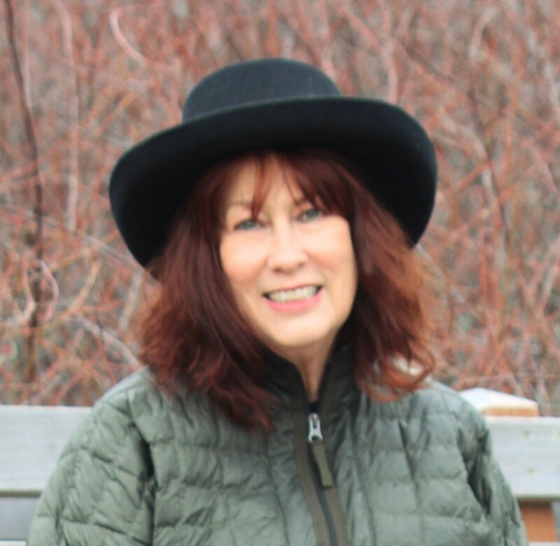 Author Marcia Aldrich smiles at the camera wearing a black hat and olive green puffer jacket.